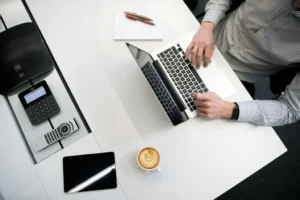 Mężczyzna pracujący na laptopie przy biurku z telefonem, tabletem i kawą, widziany z góry – usługi iod